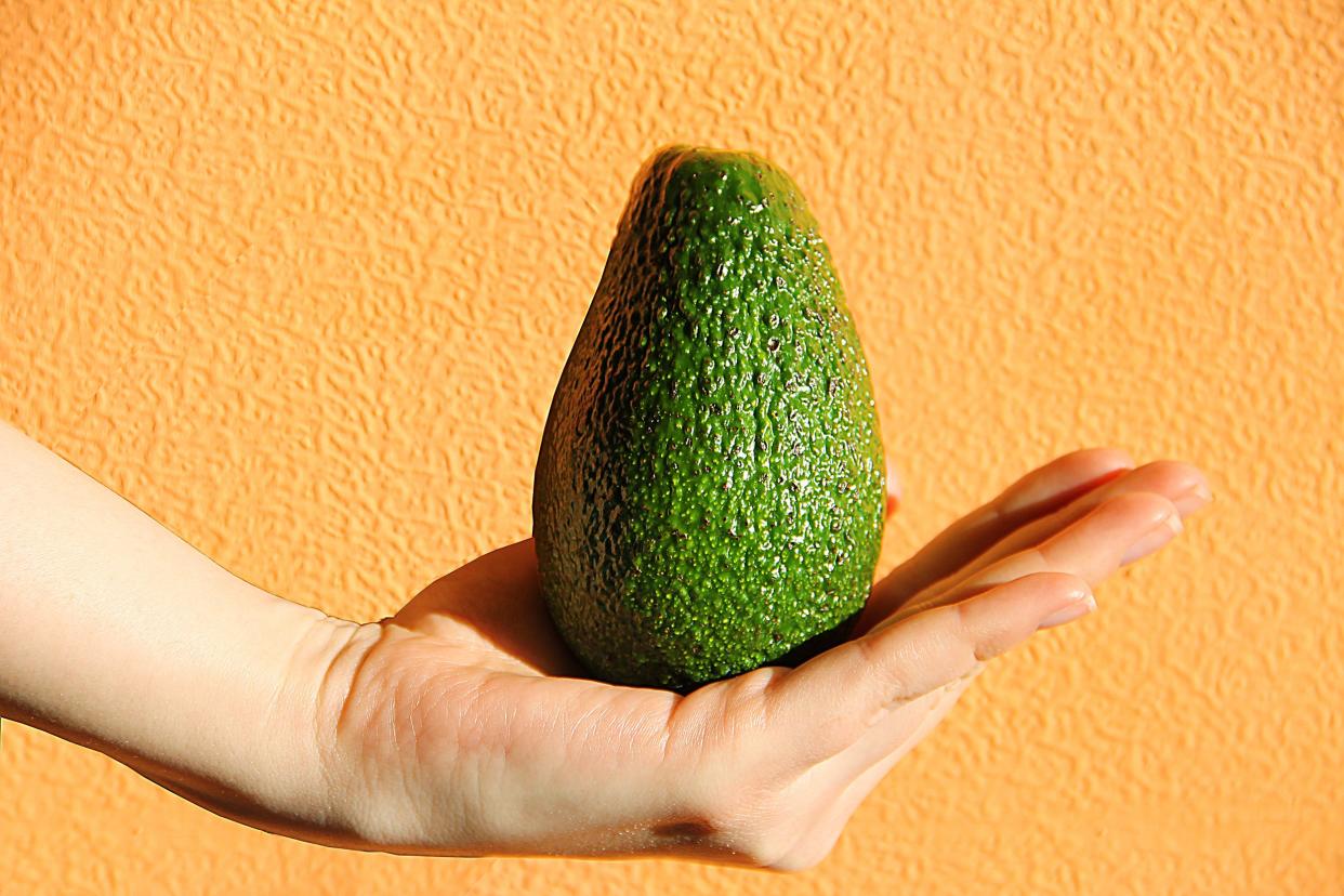 Closeup of a hand presenting an avocado against a light peachy-orange background with some texture
