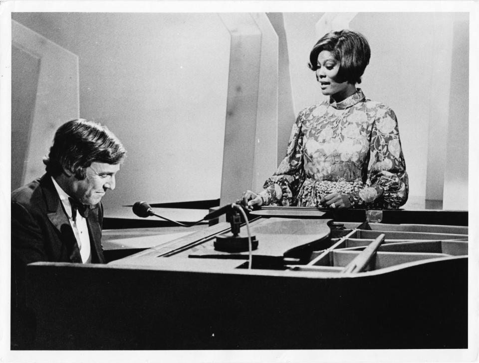 burt bacharach wearing a tuxedo and playing a piano as dionne warwick, wearing a dress, stands next to the piano and sings