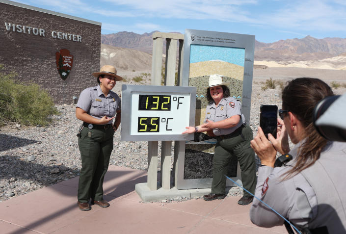 Two uniformed park rangers pose for a photo near a visitor center sign against a desert landscape while a third ranger holds a cell phone pointed at them next to a number sign that reads: 132F 55C