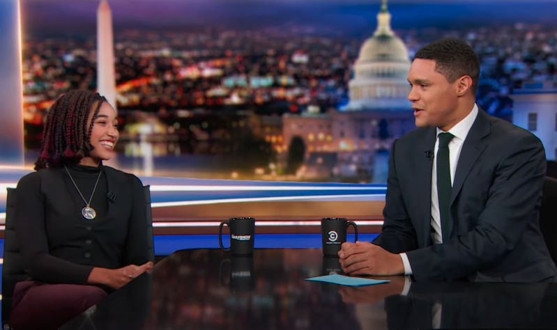 Amandla Stenberg and Trevor Noah on a late-night talk show set, talking with a cityscape background and Capitol building visible