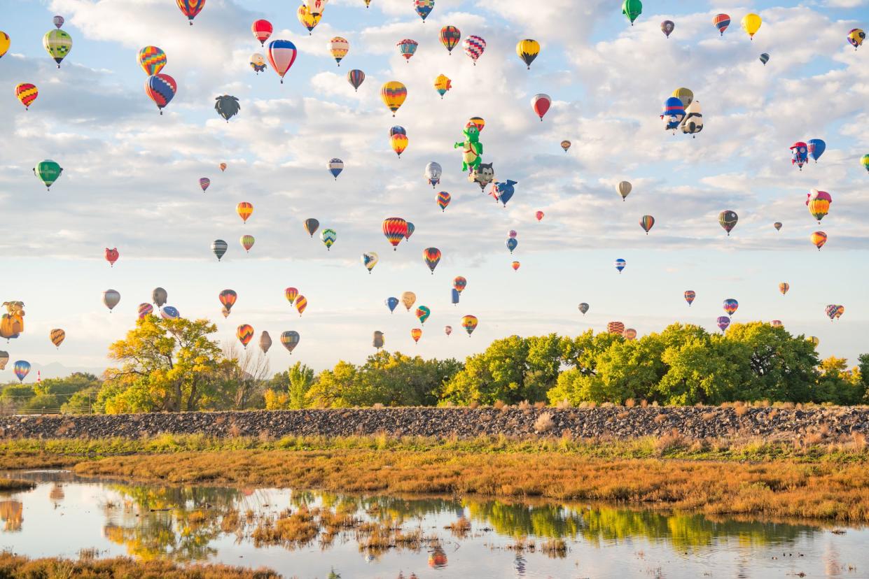 Colorful array of hot air balloons in the air during Balloon Fiesta, Albuquerque, New Mexico, creek and trees in the foreground with a sky of hot air balloons