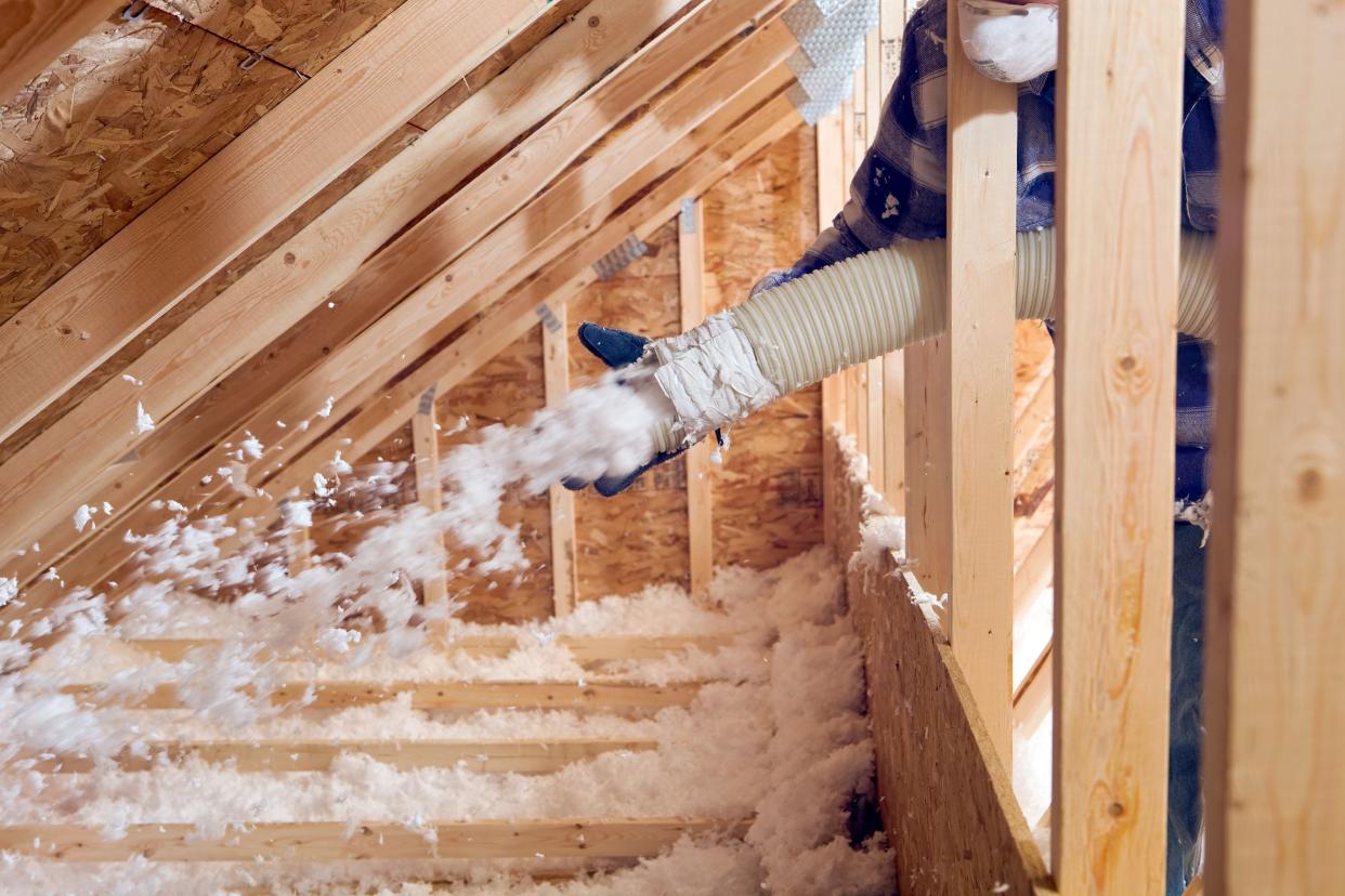 worker spraying blown fiberglass insulation between attic trusses