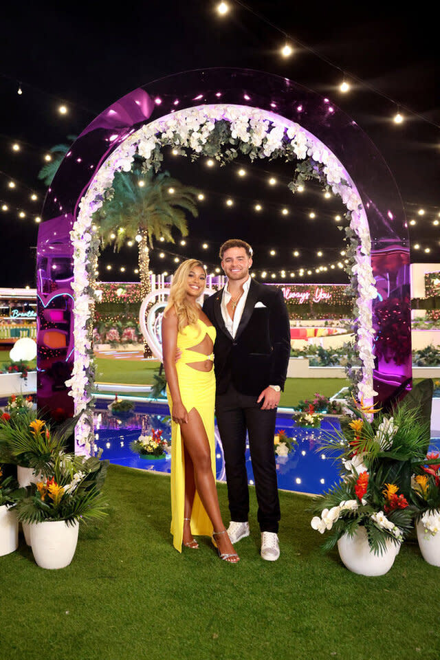 Hannah Wright wearing a yellow gown and Marco Donatelli wearing a black suit together in an outdoor decorated space.