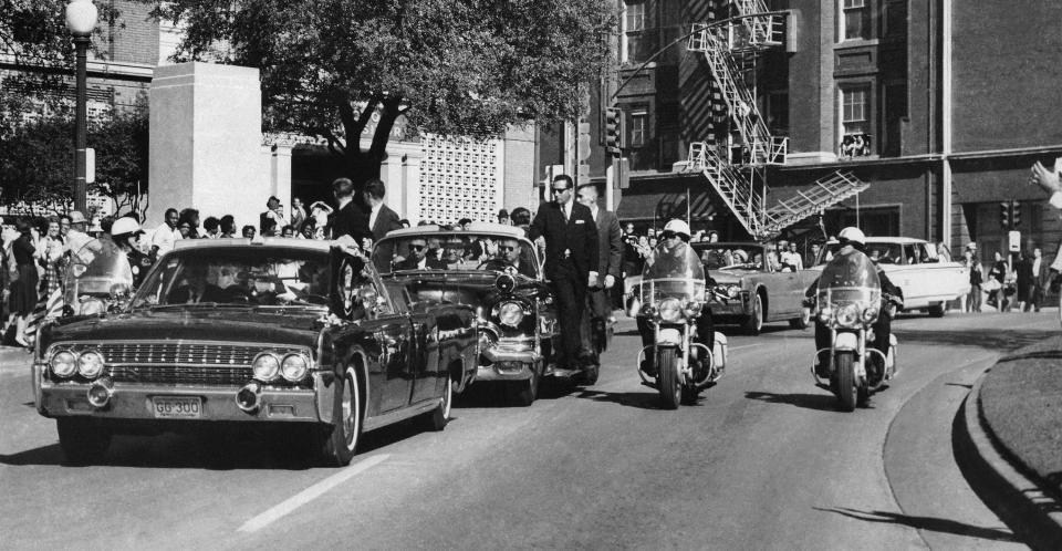 FILE - Seen through the foreground convertible's windshield, President John F. Kennedy's hand reaches toward his head within seconds of being fatally shot as first lady Jacqueline Kennedy holds his forearm as the motorcade proceeds along Elm Street past the Texas School Book Depository in Dallas, on Nov. 22, 1963. Some of the last surviving witnesses to the events surrounding the assassination of Kennedy are among those sharing their stories as the nation marks the 60th anniversary. Associated Press reporter Peggy Simpson said she rushed to the scene that day, staying with police as they converged on the Texas School Book Depository,(AP Photo/James W. "Ike" Altgens, File)