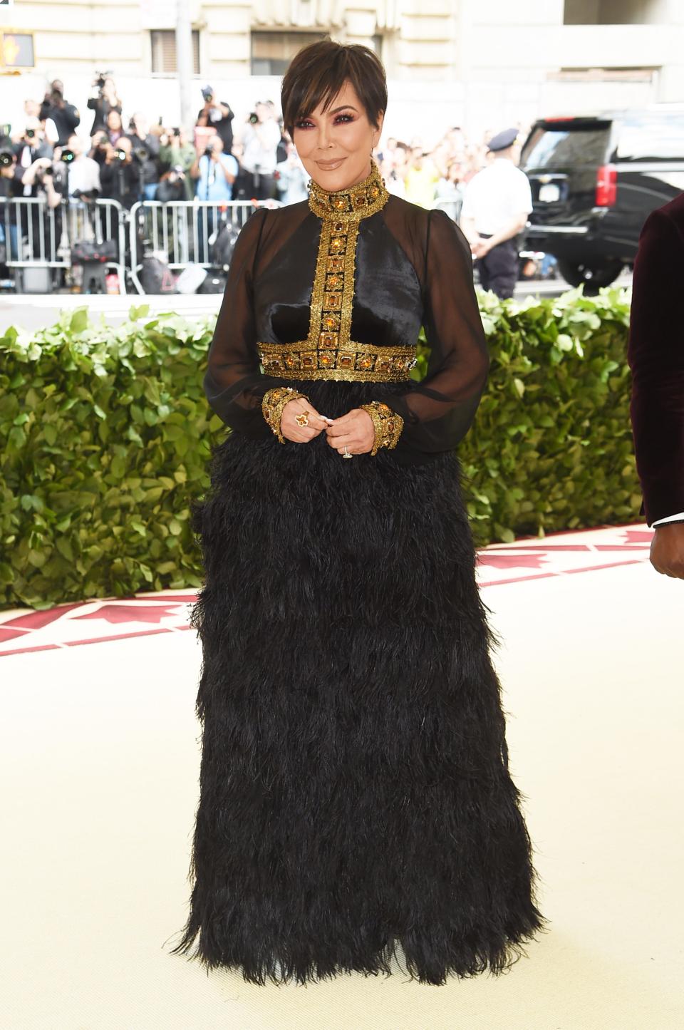 Kris Jenner at Met Gala 2018, wearing a floor-length black gown with a feathered skirt and gold embellishment on the cuffs and neck.