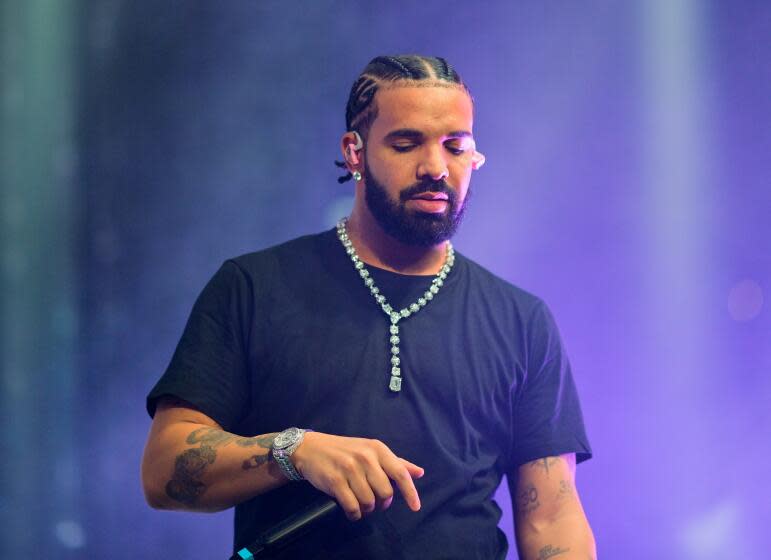 Man, Drake, hair in cornrows, on stage wearing black t shirt and diamond chain, holding mic along his chest, looking down