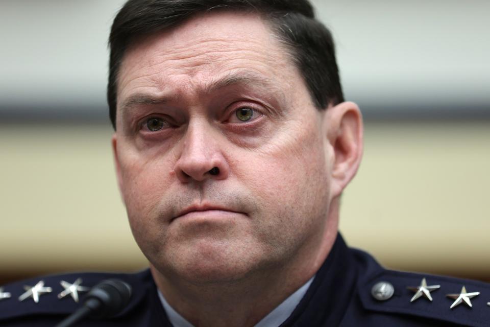 a man in a black military dress outfit sits in a senate chamber behind microphones