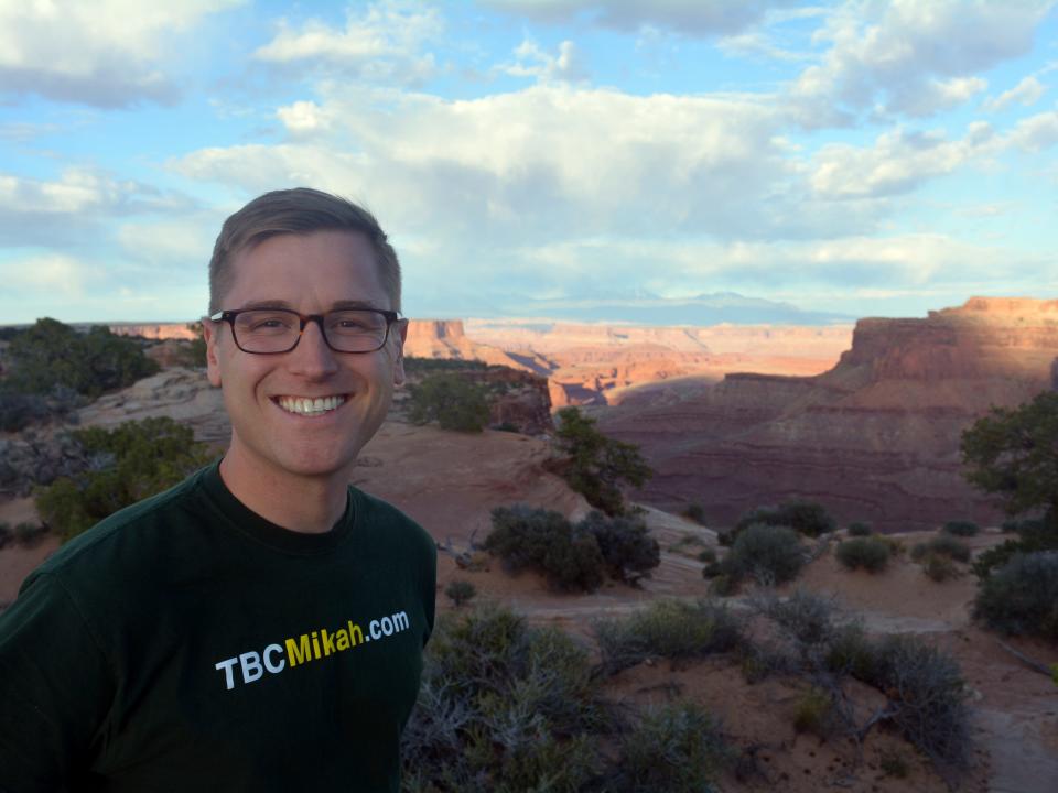 Mikah Meyer smiling on the left with canyons in the background and cloudy blue skies.