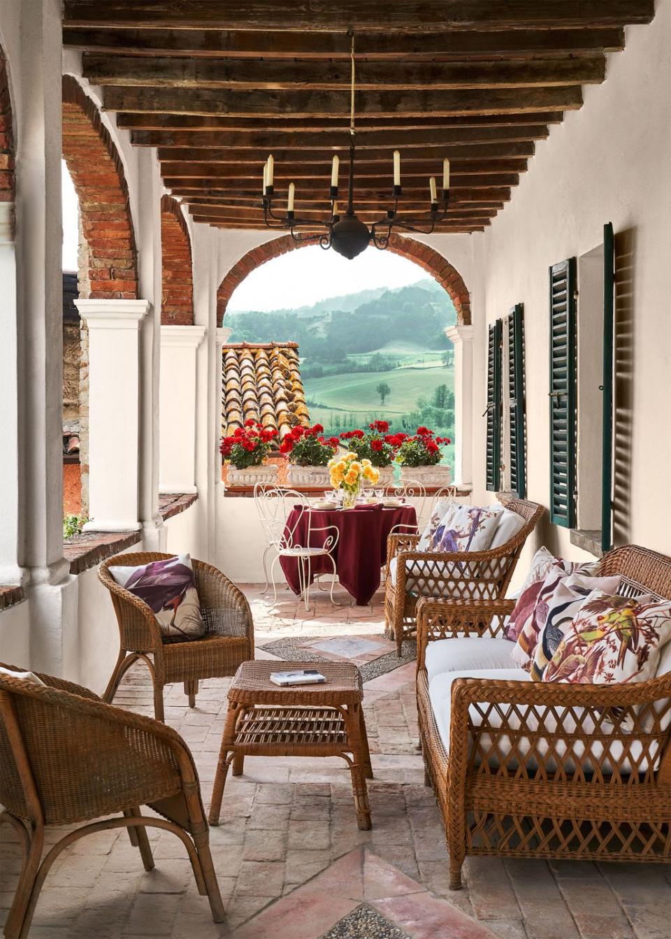 a loggia with arched brick windows, worn terra cotta floor with chairs and tables, rattan sofa, chairs, and cocktail table, a round table with cloth with curved back metal chairs, hanging metal candleholder, view of hills