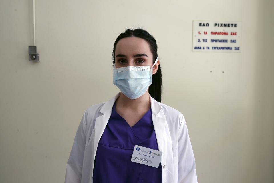 In this photo taken on Friday, May 8, 2020, medical student Anna Karagiannakou poses for a photo at the entrance of the COVID-19 Clinic at Sotiria Hospital in Athens. Greece's main hospital for the treatment of COVID-19 is also the focus of a hands-on training program for dozens of medical students who volunteered to relieve hard-pressed doctors from their simpler duties while gaining a close peek at the front lines of a struggle unmatched in modern medical history. The sign below, placed above a box that has since been removed, reads ''This is where you leave your complaints, your suggestions and your congratulations too.'' (AP Photo/Thanassis Stavrakis)