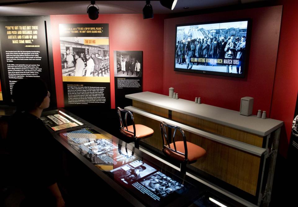 a woman looks at museum exhibit consisting with black displays of text and a television mounted on a red wall, and a diner counter with chairs underneath the television