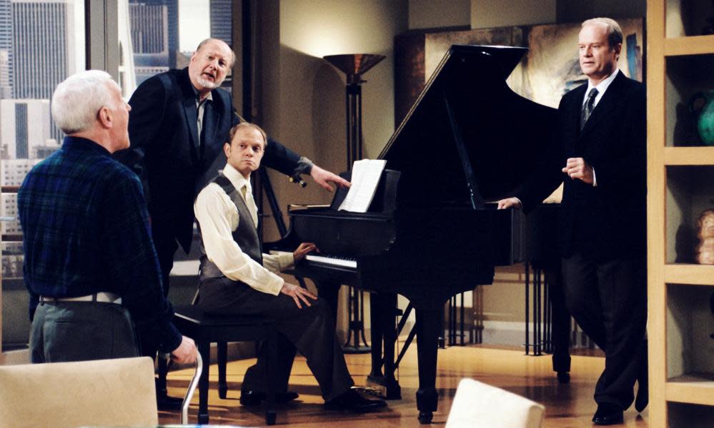 David Ogden Stiers, standing beside a piano, in the ‘Fathers and Sons’ episode of Frasier, with John Mahoney, David Hyde Pierce and Kelsey Grammer