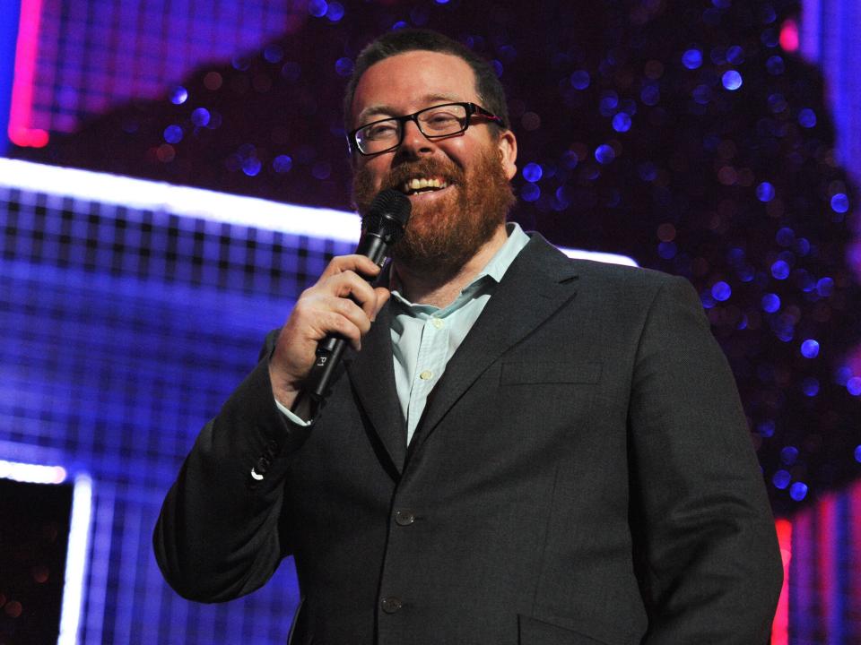 comedian frankie boyle in a button up and blazer onstage at wembley arena, smiling and holidng a microphone up to his mouth