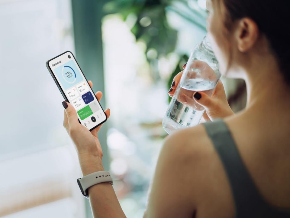 A woman checking a smart phone fitness app after a workout, drinking a glass of water, pictured from over her left shoulder
