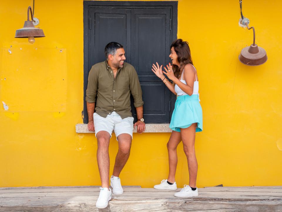 A couple talking in front of a yellow wall with a black covered window behind them.