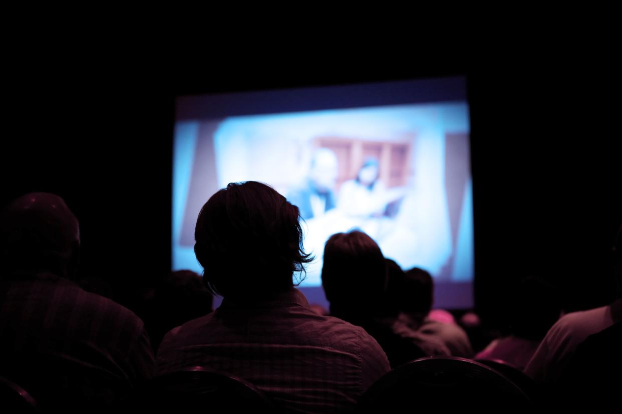 An unknown number of people sitting in a dark theater.  There is a projector screen in front of them with a man and woman at a conference.