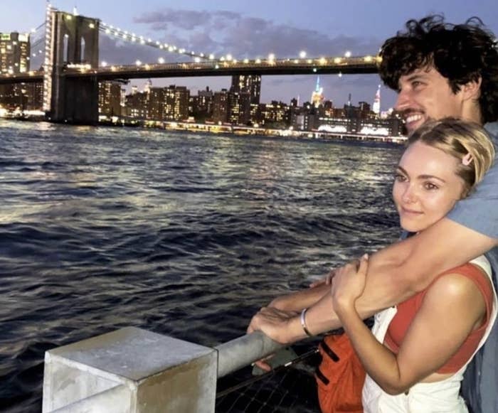 Trevor puts his arms around AnnaSophia as they look over a fence at a lit up bridge