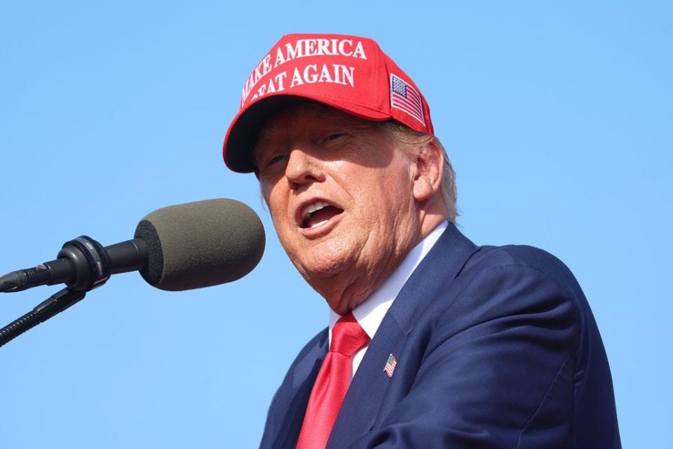 Trump speaking at a microphone while wearing a MAGA hat.