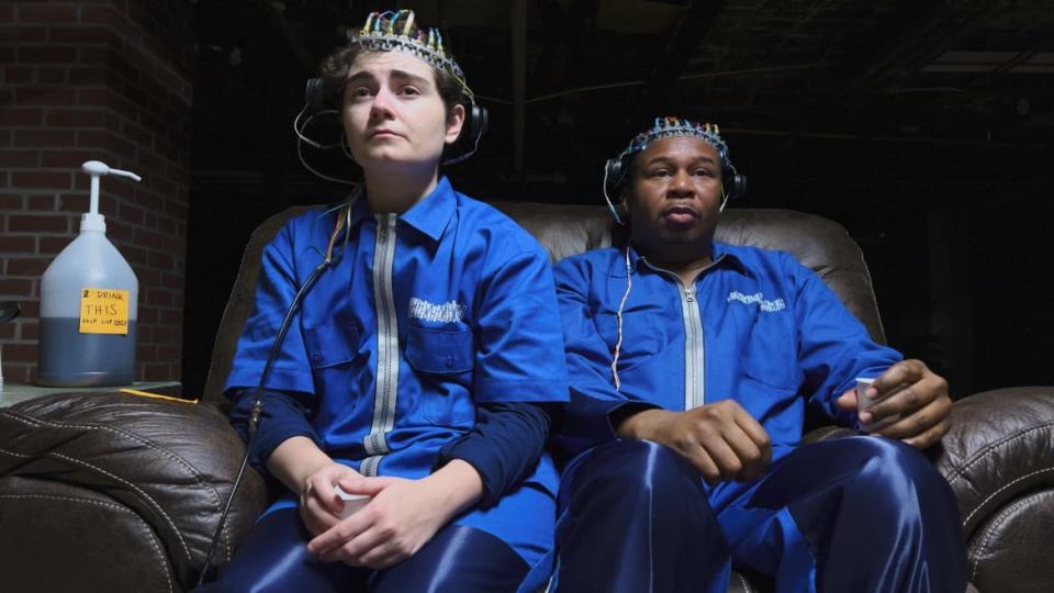 Two people sit in an armchair together, both with a wire helmet on their heads.