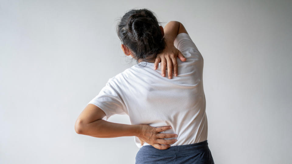 A woman in a white t-shirt holding her hand between her shoulder blades and the other on her back