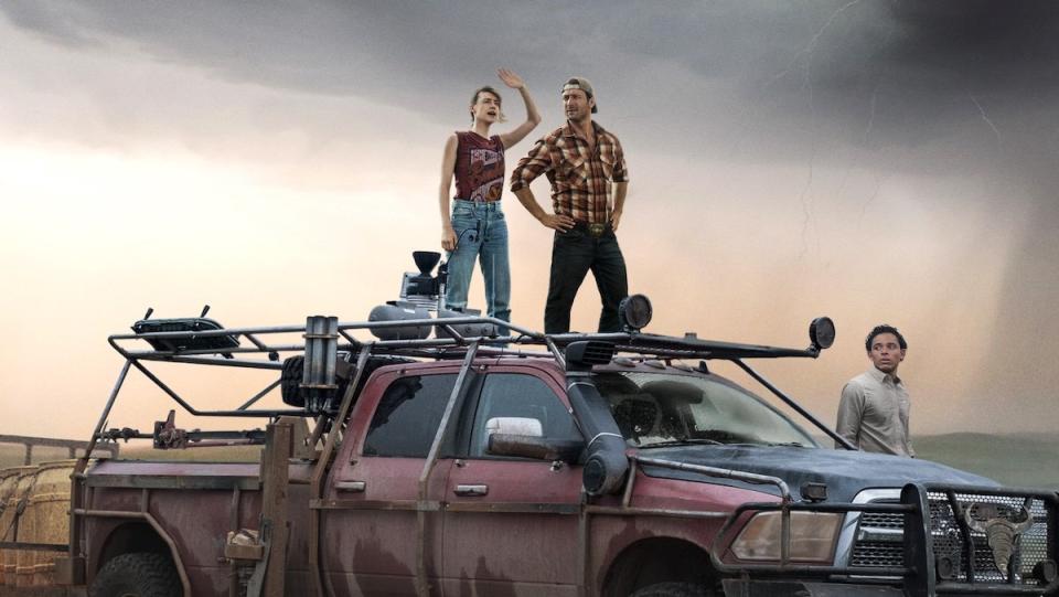 Two people stand on a red pickup truck while a man stands near the front all out in an open field with a stormy sky in a poster for Twisters
