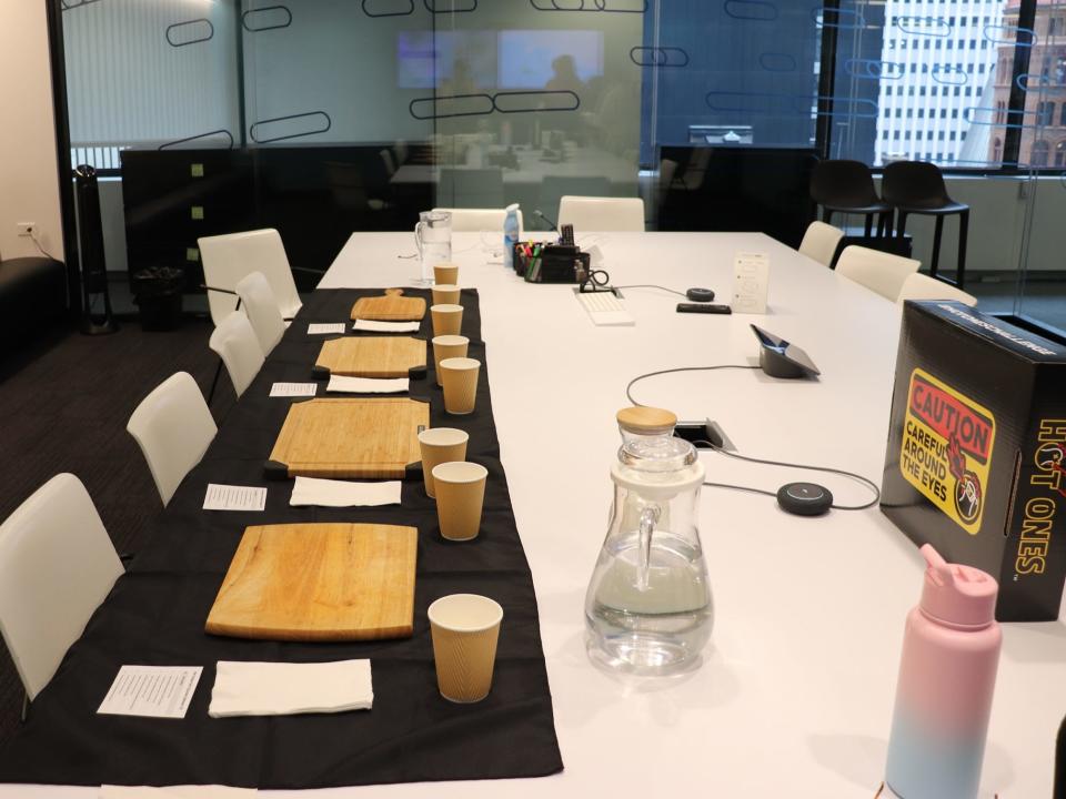 A conference room table set up with black tablecloths, wooden cutting boards, napkins, and cups.