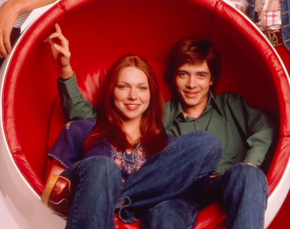 Donna Pinciotti and Eric Forman characters sitting in a large red bowl chair, in casual 70s attire, from "That '70s Show."