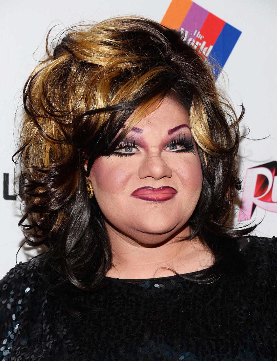 A person with voluminous hair and dramatic makeup smiles for the camera. They are wearing a sparkly black top