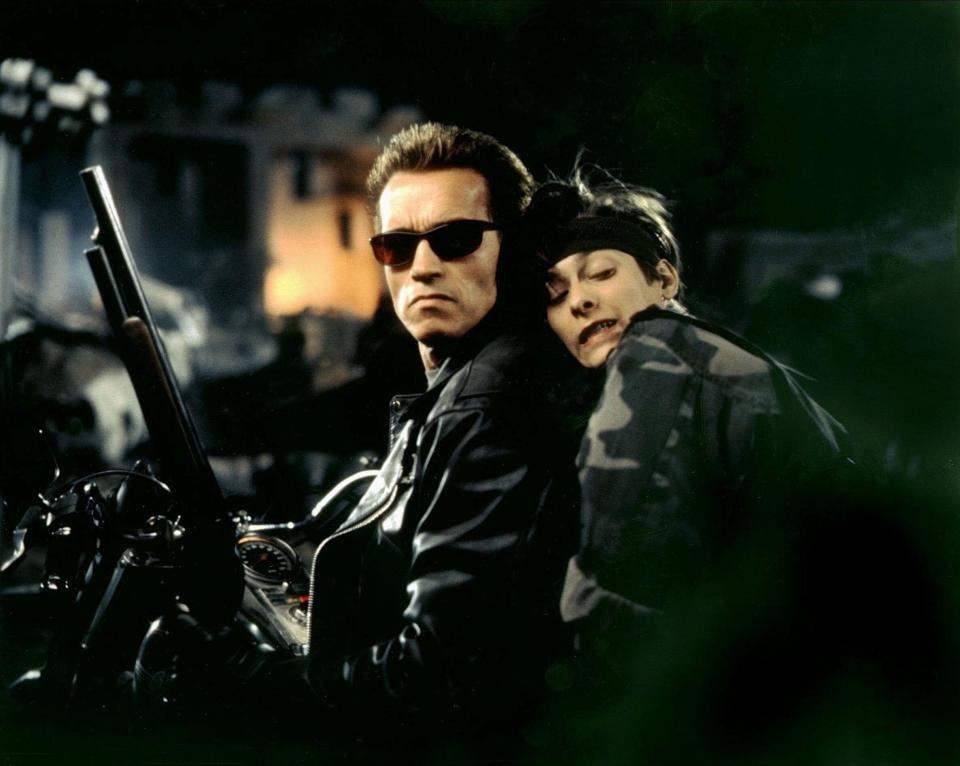 A young child in a camouflage jacket sits behind leather jacket–wearing Schwarzenegger as the T-800 on a motorcycle