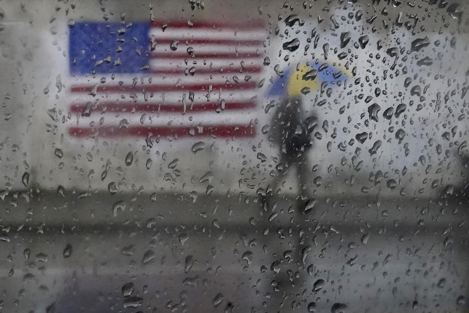 A pedestrian carries an umbrella while walking past a painting of the American flag in San Francisco, Wednesday, Jan. 11, 2023. Storm-ravaged California is scrambling to clean up and repair widespread damage. (AP Photo/Jeff Chiu)