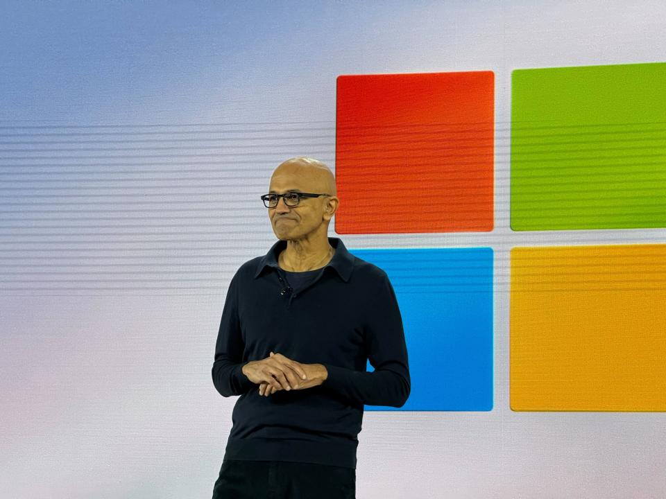 Satya Nadella on stage with the Windows logo on a screen behind him.