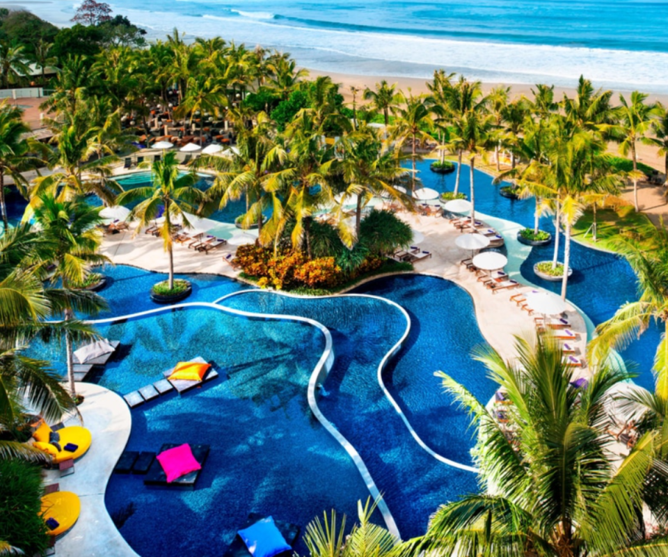 A birdseye view of the W Bali's turquoise pool in the shape of sloping rice fields with palm trees surrounding the pools and the ocean in the right side of the background.