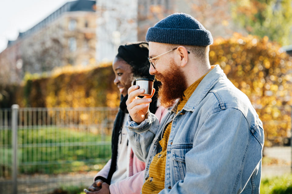 A father sitting next to his wife and drinking some coffee from a flask while spending the afternoon at the park with his family.