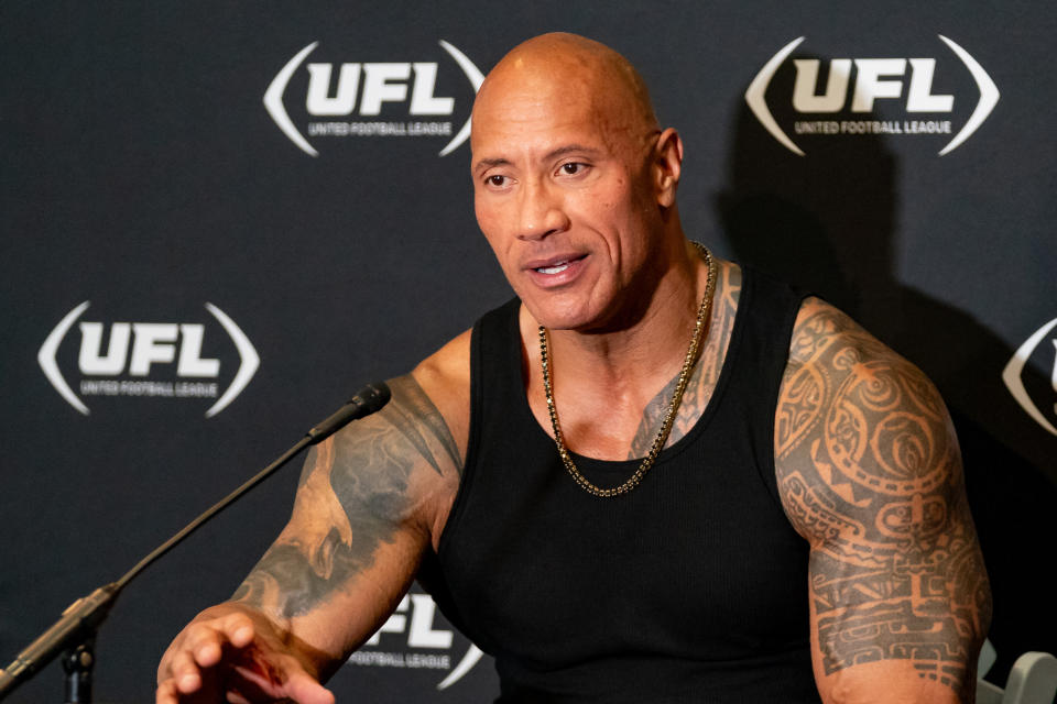 Dwayne Johnson speaks at a UFL event, wearing a black tank top, with visible tattoos