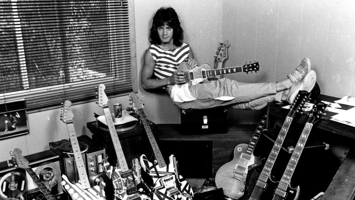  Eddie Van Halen sitting down holding a guitar with lots of other guitars on the floor. 