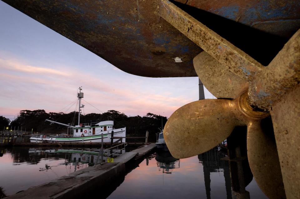 A green and white boat is seen from a vantage point below the exposed propeller of a larger vessel.