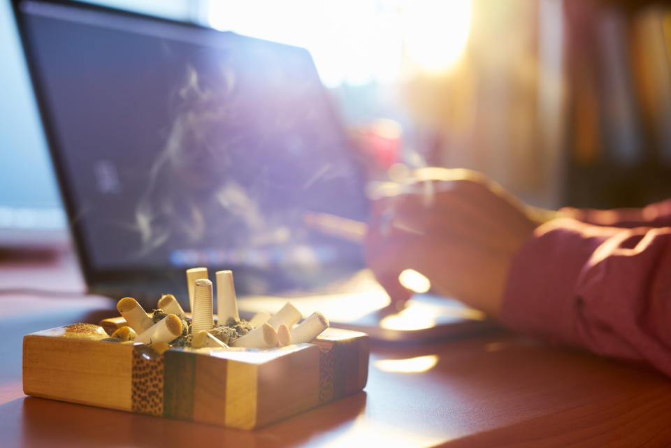 a person looking at their laptop with a cigarette in their hand and a filled ash tray next to them