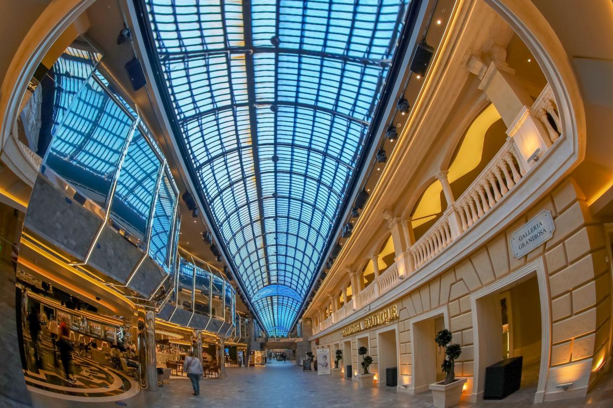 Luxurious shopping area inside the MSC Grandiosa cruise ship, blue sky through the ceiling windows, high-end shops on both sides