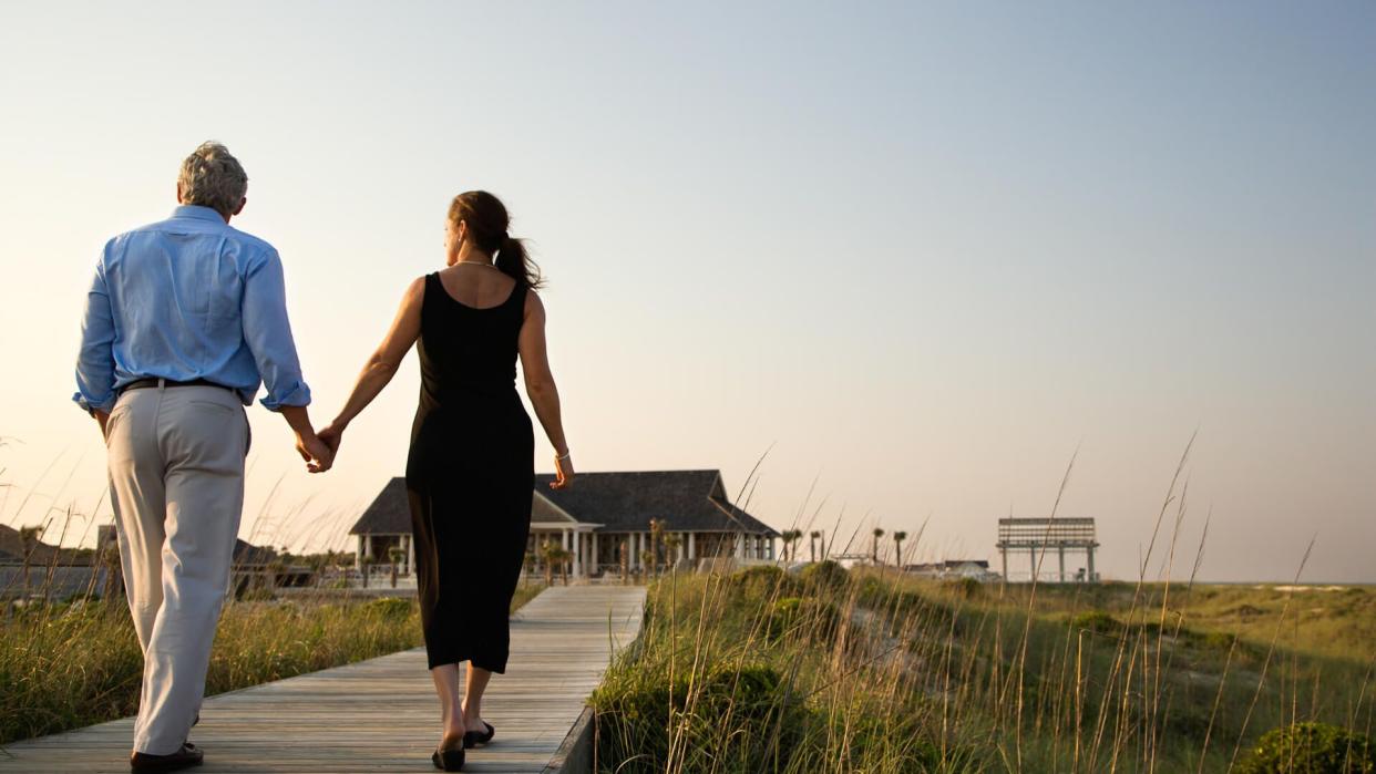 Couple walk hand in hand on a boardwalk towards a beach pavilion.