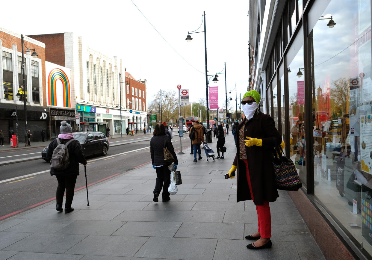 A woman wearing a scarf and plastic gloves as a precautionary measure for Covid-19, waits for a bus in the high street in Brixton, south London, as the UK continues in lockdown to help curb the spread of the coronavirus. Picture date: Monday April 6, 2020. 