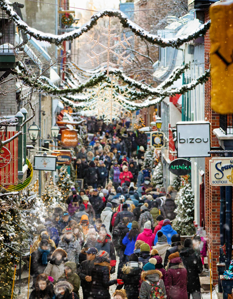 Crowded, festive pedestrian street decorated for winter holidays with hanging garlands and a large snowflake. People are shopping, chatting, and enjoying the day