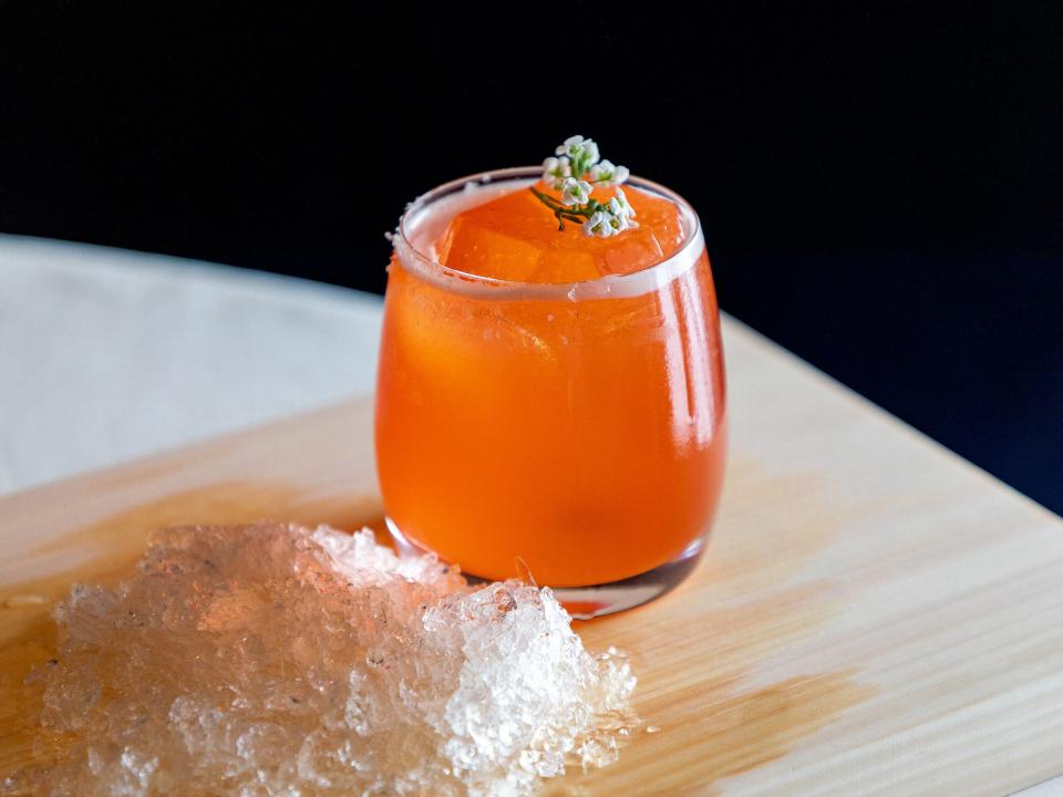 Orange beverage from Providence with white floral garnish on top of a wooden surface, with crushed ice on the side