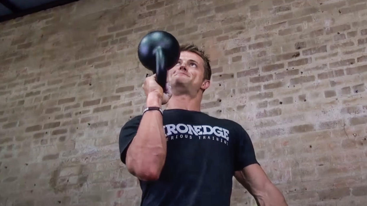 Man peforming a bottoms-up kettlebell press standing against a brick wall. 
