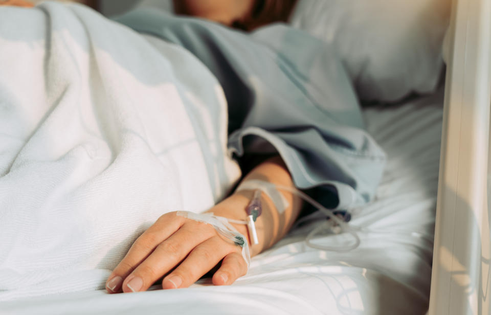 A person with an IV in their hand lies in a hospital bed, covered with a blanket