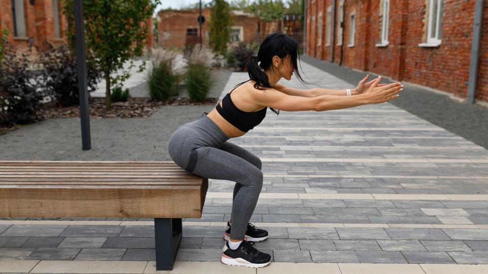 Woman sitting on a bench outside with arms extended in front of her during squat