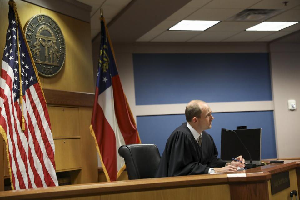 A man in a black robe sits behind a desk, flanked by the U.S. flag and the Georgia state flag.