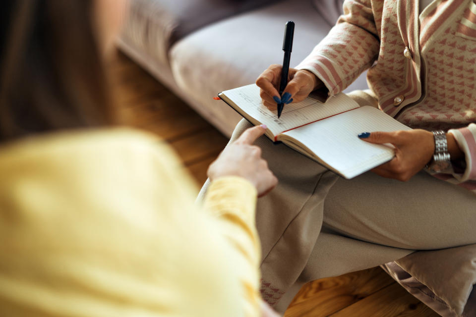 Two people are sitting on a couch. One is writing in a notebook while the other is pointing at it. Both faces are not visible.