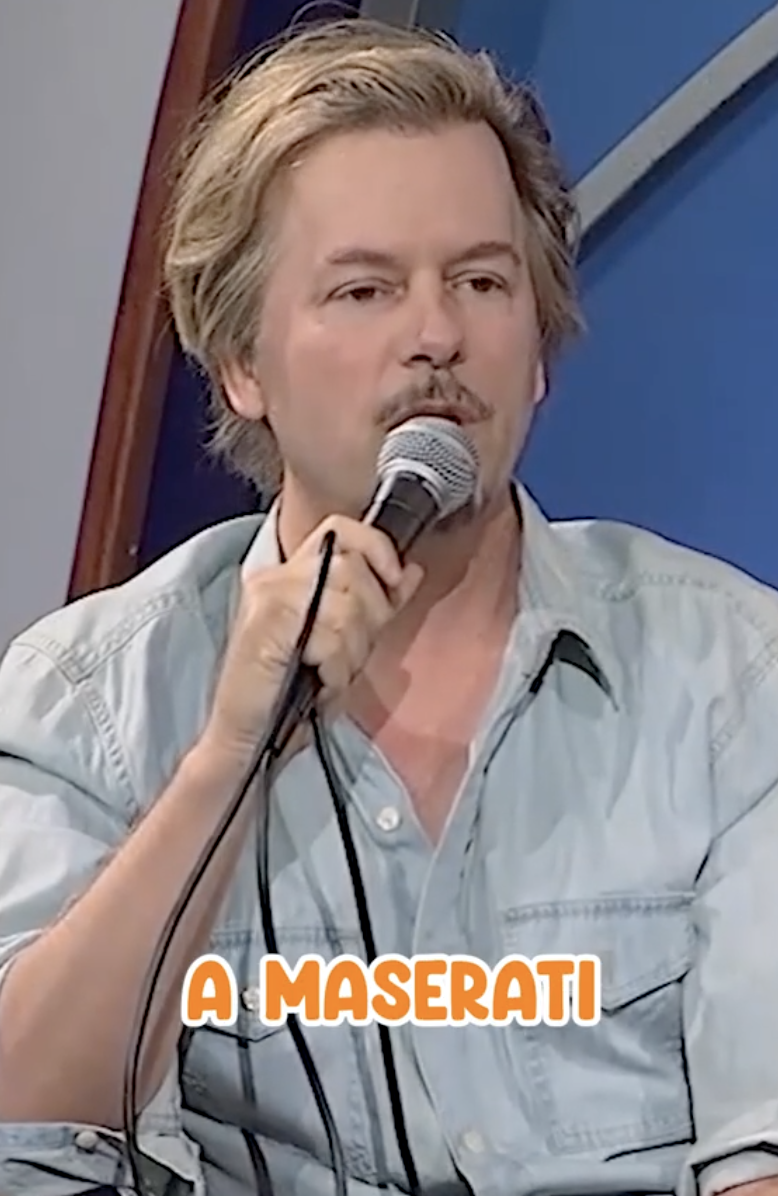 David Spade speaks into a microphone while wearing a casual button-up shirt. Text at the bottom of the image reads 