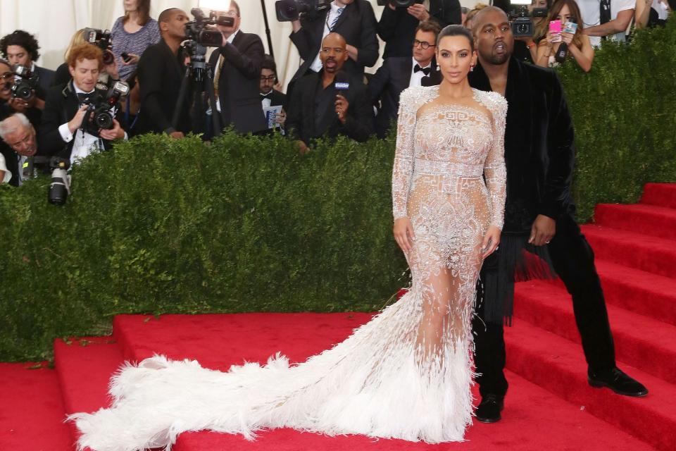 Kim Kardashian poses for photos with then-husband Kanye West on the Met Gala red carpet. She's wearing a nude gown with silver embellishments and a white feathered skirt while he's wearing a black suit with a velvet jacket.