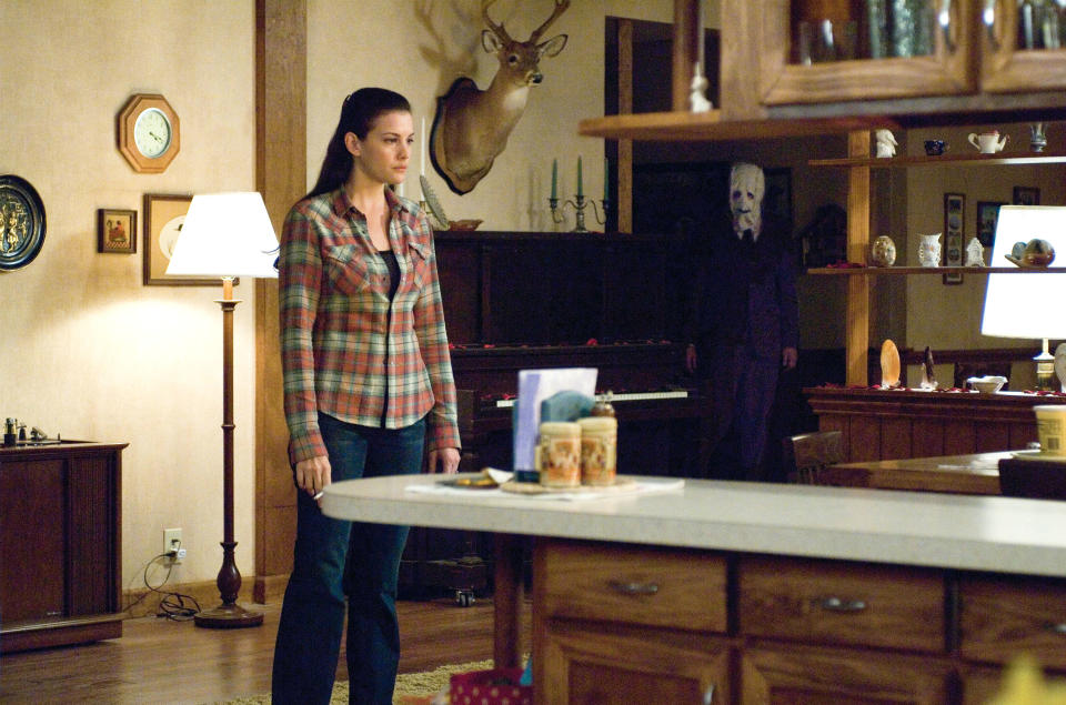 Liv Tyler standing in a room with a man in a scary mask off to the side and behind her, facing her